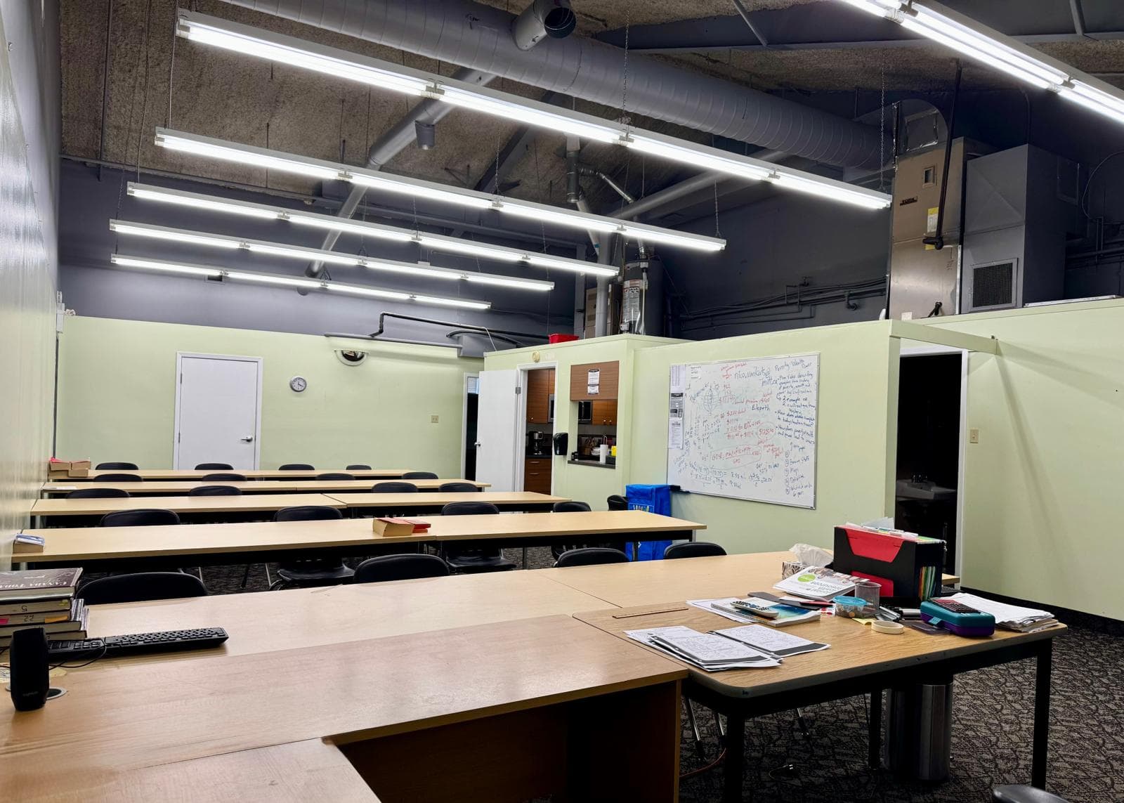 A long training room with rows of tables and chairs under exposed ceiling ducts and strip lighting, pale green partition walls, and a whiteboard at the far end