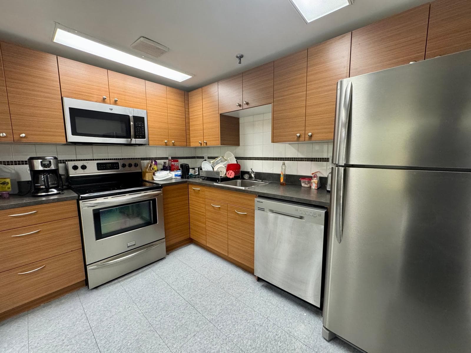A kitchen with wood-fronted cabinets, a stainless fridge, dishwasher and over-range microwave, and a coffee maker and kettle on the counter