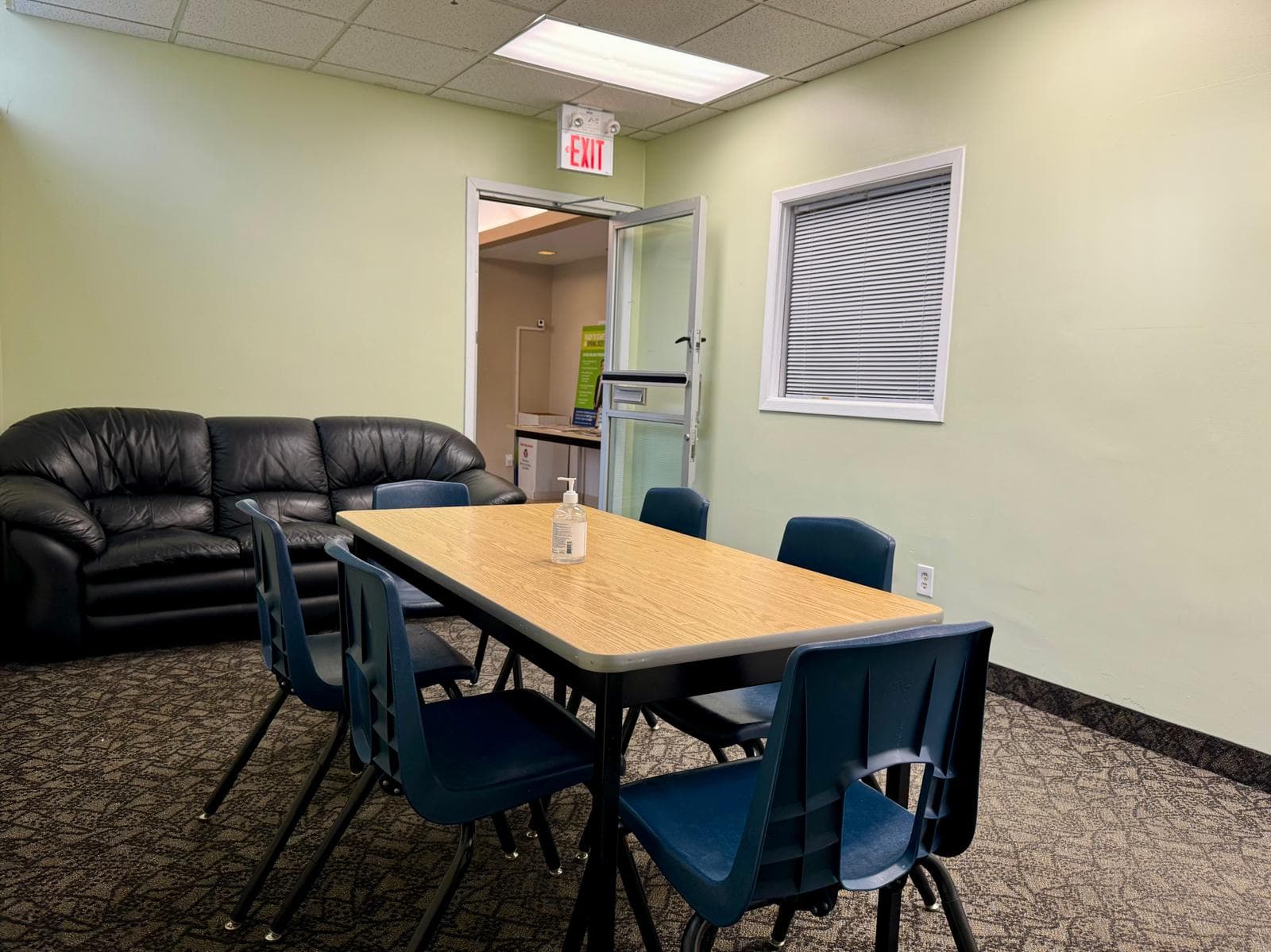 A meeting room with a wooden table and blue stacking chairs, a black leather sofa against one wall, and a window with closed blinds