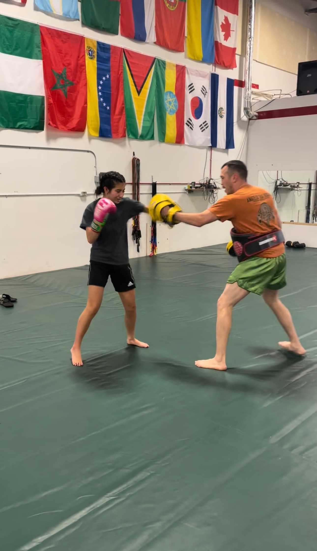 A young person in gloves throwing a punch at focus mitts held by a coach on a gym mat, with national flags hanging along the wall behind them