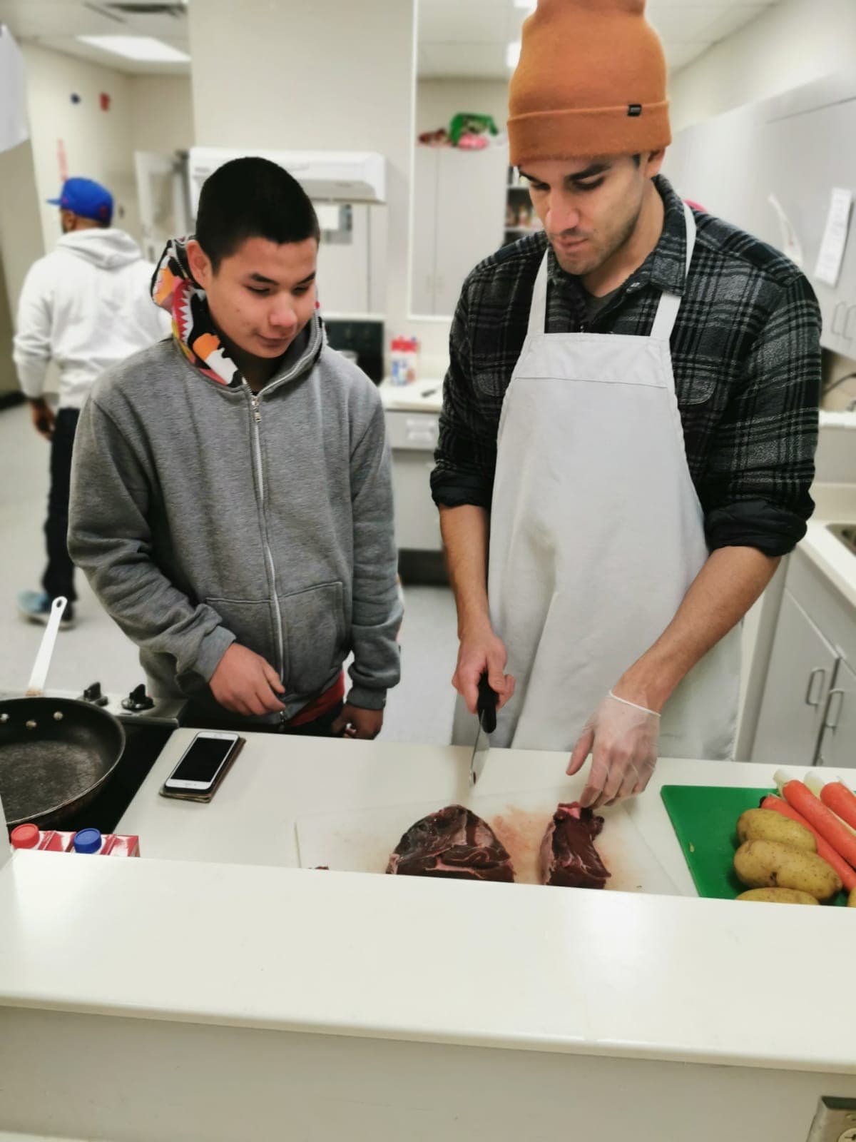 A mentor in an apron slicing meat at a kitchen counter while a young person watches
